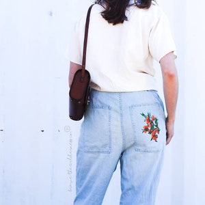 May include: A person wearing a white shirt and light blue jeans with a floral embroidery on the back pocket. The person is carrying a brown leather shoulder bag.