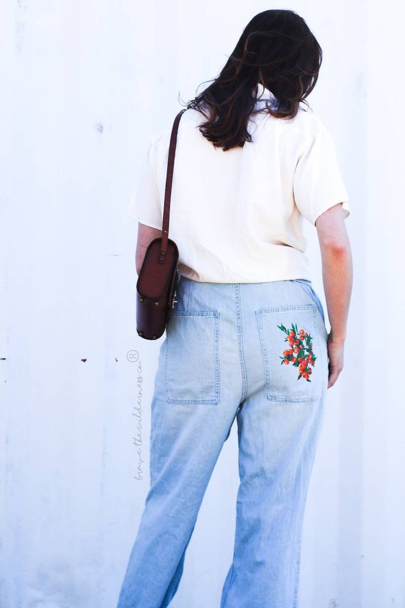 May include: A person wearing a white shirt and light blue jeans with a floral embroidery on the back pocket. The person is carrying a brown leather shoulder bag.