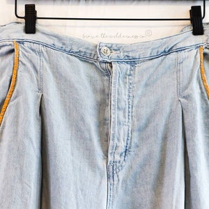 May include: Light blue denim pants with a brown stitched detail along the sides. The pants have a button fly and a waistband with the text "browse the wilderness co." printed on it.