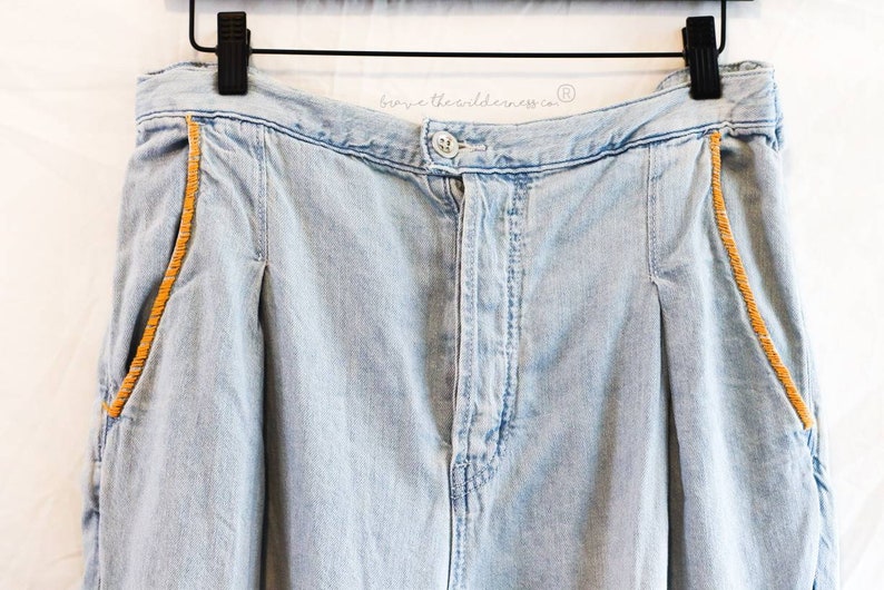 May include: Light blue denim pants with a brown stitched detail along the sides. The pants have a button fly and a waistband with the text "browse the wilderness co." printed on it.
