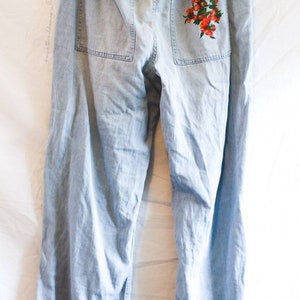 May include: Light blue denim pants with a floral embroidery design on the back pocket. The embroidery features red flowers and green leaves.