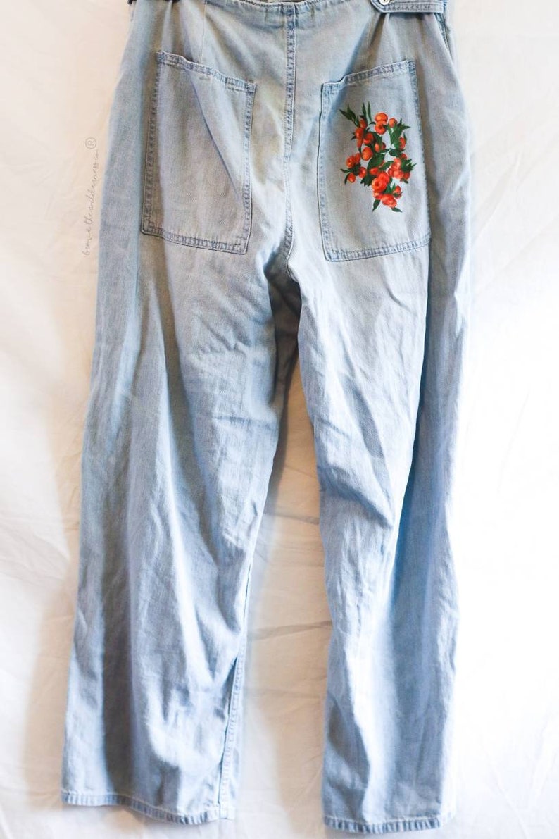May include: Light blue denim pants with a floral embroidery design on the back pocket. The embroidery features red flowers and green leaves.