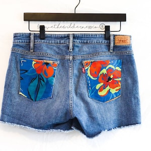 May include: Blue denim shorts with frayed edges. The back pockets are decorated with colorful floral fabric patches.