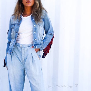 May include: A light blue denim jacket with frayed edges and red and blue fringe detail on the sleeves. The jacket is worn over a white t-shirt and light blue denim pants.
