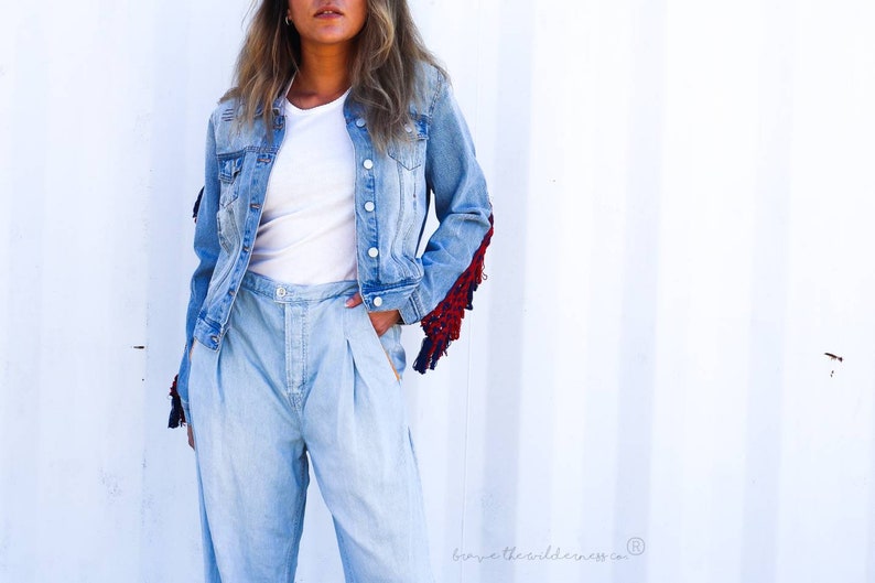 May include: A light blue denim jacket with frayed edges and red and blue fringe detail on the sleeves. The jacket is worn over a white t-shirt and light blue denim pants.