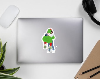 May include: A sticker featuring a cartoon illustration of two figures embracing. One figure is in a green costume with a red hat, and the other is wearing a red and white baseball jersey, blue jeans, and sneakers. The sticker is on a laptop.