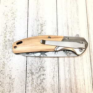 Op de afbeelding: Een vouwmes met een houten handvat en een roestvrijstalen mes. Het handvat is lichtbruin met een houtnerfpatroon. Het mes is zilverkleurig met een zwart-wit camouflagepatroon.