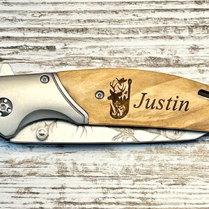 Op de afbeelding: Een gepersonaliseerd zakmes met een houten handvat. Het handvat is voorzien van een lasergegraveerd beeld van een hert met een vislood en de naam "Justin".