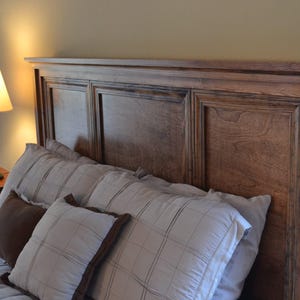 May include: A wooden headboard with a paneled design. The headboard is brown and has a distressed finish. The headboard is made of wood and has a traditional style. The headboard is decorated with pillows in a variety of colors and patterns.