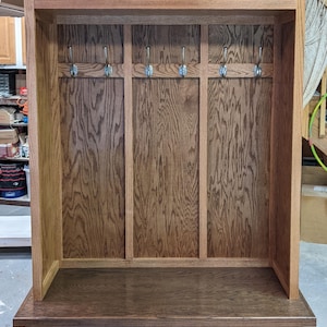 May include: A wooden entryway bench with a coat rack and storage compartments. The bench has a dark brown stain and features two drawers with a raised panel design. The coat rack has five hooks.
