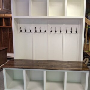 May include: White wooden entryway bench with a dark brown wood top, three storage cubbies, and a top shelf with three cubbies. The bench has a row of hooks for hanging coats or bags.