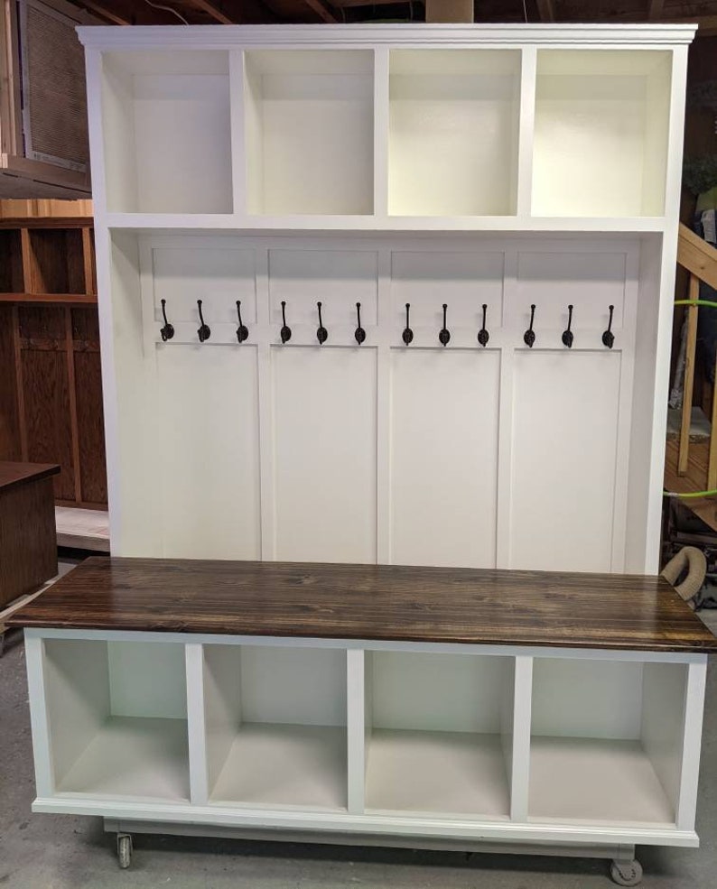 May include: White wooden entryway bench with a dark brown wood top, three storage cubbies, and a top shelf with three cubbies. The bench has a row of hooks for hanging coats or bags.