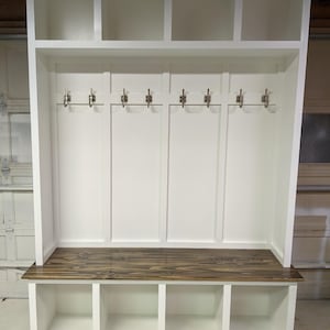 May include: White wooden entryway bench with a dark wood top, three storage cubbies, and a top shelf with three open cubbies. The bench has six metal hooks for hanging coats or bags.
