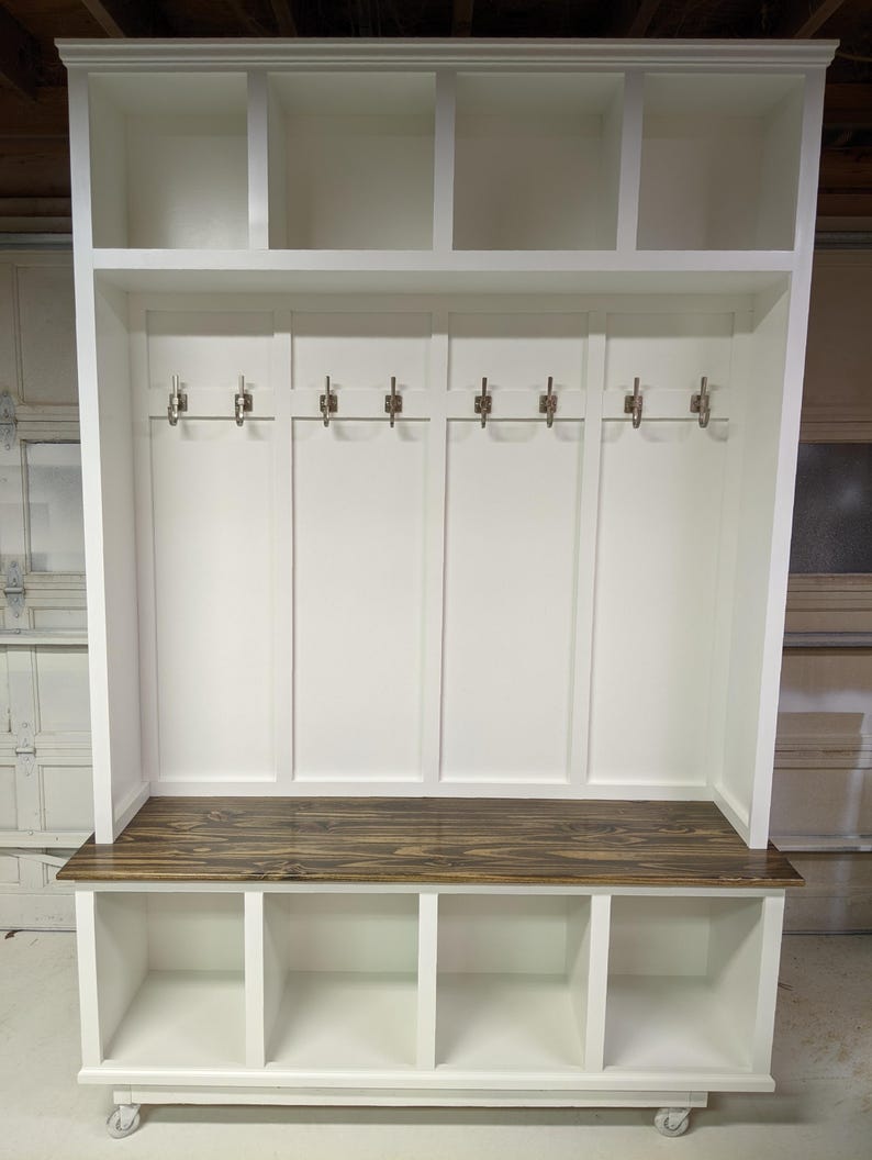 May include: White wooden entryway bench with a dark wood top, three storage cubbies, and a top shelf with three open cubbies. The bench has six metal hooks for hanging coats or bags.