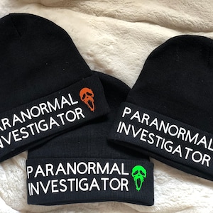May include: Three black knit beanies with white text that reads "Paranormal Investigator" and a ghost face graphic in orange, pink, and green.