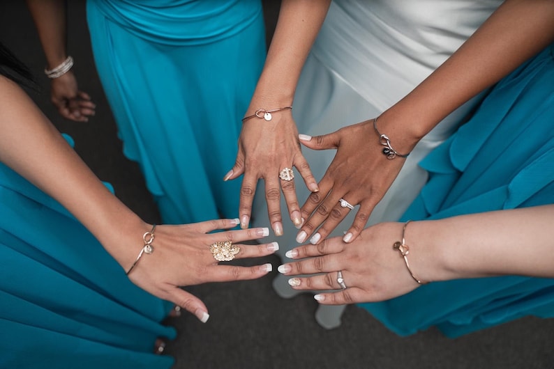 May include: Four hands with white manicured nails are shown wearing rings and bracelets. The hands are wearing gold and silver jewelry. The hands are against a background of blue fabric.