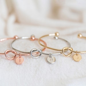 May include: Three gold knot bracelets with initial charms. The bracelets are in rose gold, silver, and gold.
