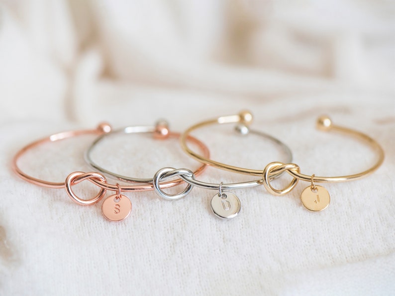 May include: Three gold knot bracelets with initial charms. The bracelets are in rose gold, silver, and gold.