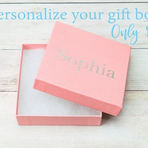 Puede incluir: Una caja de regalo rosa con una tapa blanca y una inscripci&oacute;n blanca que dice "Sophia". La caja est&aacute; abierta y la tapa est&aacute; apoyada en el lado derecho. La caja est&aacute; sobre una superficie de madera blanca.