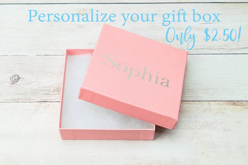 Puede incluir: Una caja de regalo rosa con una tapa blanca y una inscripci&oacute;n blanca que dice "Sophia". La caja est&aacute; abierta y la tapa est&aacute; apoyada en el lado derecho. La caja est&aacute; sobre una superficie de madera blanca.