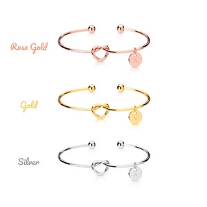 May include: Three gold knot bangle bracelets with a small charm hanging from each. The charms are round and have a letter on them. The bracelets are shown in rose gold, gold, and silver.