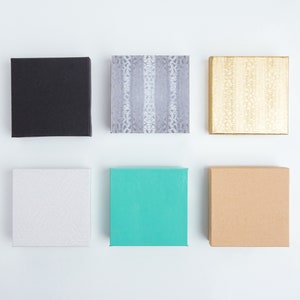 Puede incluir: Seis peque&ntilde;as cajas cuadradas de diferentes colores y patrones. Las cajas est&aacute;n dispuestas en dos filas de tres. La fila superior presenta una caja negra, una caja gris con un patr&oacute;n blanco y una caja dorada con un patr&oacute;n blanco. La fila inferior presenta una caja blanca con un patr&oacute;n sutil, una caja turquesa y una caja marr&oacute;n.