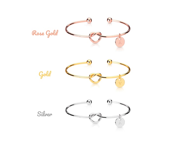 Puede incluir: Tres pulseras de nudo de oro con un peque&ntilde;o dije colgando de cada una. Los dijes son redondos y tienen una letra en ellos. Las pulseras se muestran en oro rosa, oro y plata.