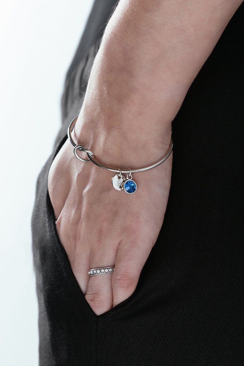 Puede incluir: Un brazalete de plata con un dise&ntilde;o de nudo y dos colgantes. Un colgante es un disco redondo de plata con una piedra blanca, y el otro es una piedra redonda azul.