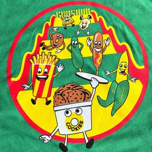 May include: A green t-shirt with a circular design featuring various food items with faces. The design includes a pizza slice, a corn dog, a pickle, french fries, a banana, and a container of cookies. The text "COOKIES" is printed on the container.