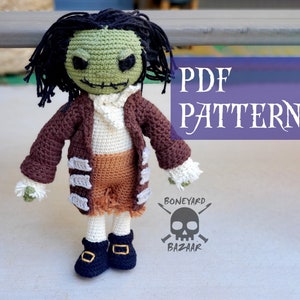 以下が含まれることがあります： 緑色のゾンビ人形の編み物パターンPDF。茶色のジャケット、白いシャツ、茶色のズボン、黒い靴を着用しています。このパターンはBoneyard Bazaarから入手できます。