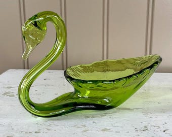 May include: A green glass swan shaped dish. The swan's neck is curved and the dish is shaped like a bowl. The dish is sitting on a white surface.