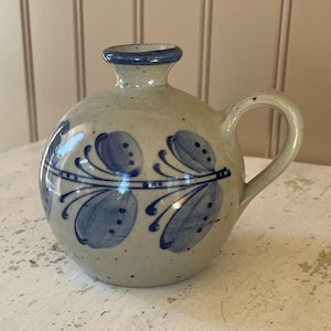 May include: A small, round, gray ceramic pitcher with a blue and white floral design. The pitcher has a handle and a spout.