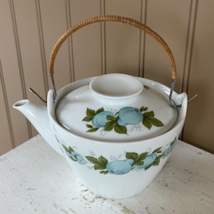 May include: A white teapot with a blue and green floral design and a woven bamboo handle. The teapot is sitting on a white wooden table.
