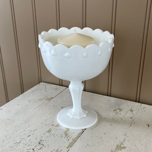 May include: White pedestal bowl with a scalloped edge and a white candle inside. The bowl is sitting on a white table with a distressed finish.