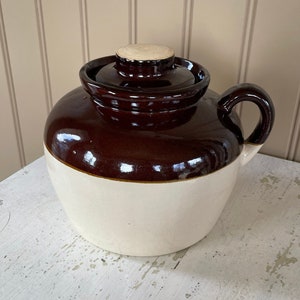 May include: A two-tone ceramic jug with a handle and a lid. The top half is a glossy dark brown, while the bottom half is a creamy white. The lid has a wooden stopper. The jug is sitting on a distressed white surface.