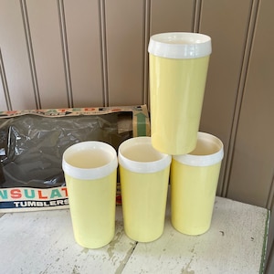 May include: Set of four yellow insulated tumblers with white lids. The tumblers are stacked on a white wooden surface. The box for the tumblers is visible in the background, with the words "Insulated Tumblers" printed on it.