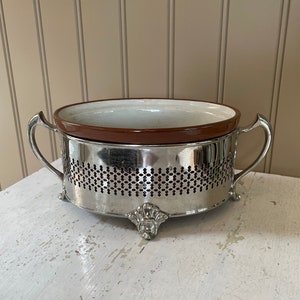 May include: A silver serving dish with a brown ceramic insert. The dish has two handles and sits on three feet.