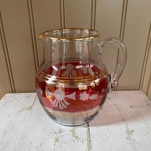 May include: A clear glass pitcher with a red and gold design. The pitcher has a wide mouth and a handle.