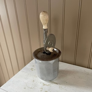 May include: A vintage silver butter churn with a wooden handle and a serrated blade. The churn is sitting on a white table.