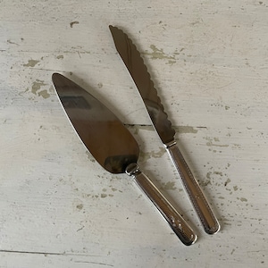 May include: Two silver cake servers, one with a serrated edge and one with a smooth edge, on a white wooden surface.