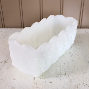 May include: A rectangular, white, vintage-style dish with a scalloped rim. The dish has a textured surface and is likely made of glass or ceramic. The sides of the dish have a floral design. The dish is empty and sits on a white surface.