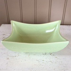 May include: A light green, rectangular ceramic dish with a curved, flared rim. The dish has a smooth, glossy finish and sits on a white, distressed wooden surface. The background features vertical, light brown wooden paneling.