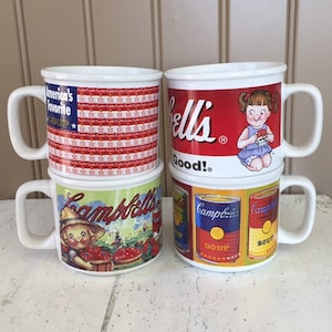 May include: Four white ceramic mugs with different Campbell's Soup designs. The mugs are stacked in pairs. One mug features the American flag with the words "America's Favorite Soup". Another mug features a girl holding a can of soup with the words "Campbell's Good!" The other two mugs feature a red and white Campbell's Soup can design.