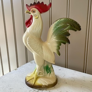 May include: A ceramic rooster figurine in cream, red, and green. The rooster has a red comb and wattle, green tail feathers, and stands on a base with green grass accents. The figurine is a decorative item.