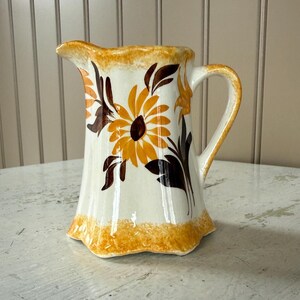 May include: A vintage ceramic pitcher with a white base, adorned with a large sunflower design in shades of orange and brown. The pitcher has a scalloped rim and base, and a curved handle. A decorative piece for the kitchen.
