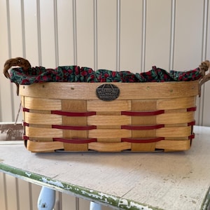 May include: A rectangular woven basket with a red and tan design. The basket has a green and red floral fabric lining the interior. The handles are made of twisted rope. A metal emblem is attached to the front of the basket.
