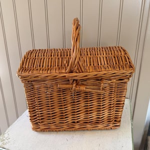 May include: A rectangular light brown wicker basket featuring a woven lid and a single upright handle. The basket is constructed with a tight weave pattern and includes a small horizontal latch mechanism on the front to secure the lid in place.