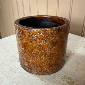 May include: A cylindrical, brown ceramic pot with a textured surface. The pot has a dark interior and a wide, rounded rim. The exterior features a mottled, aged appearance, suggesting a vintage or rustic aesthetic. The pot is suitable for plants or decorative use.