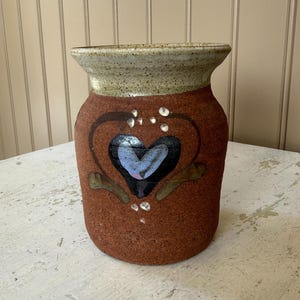 May include: A cylindrical ceramic vase with a speckled beige rim and a textured reddish-brown body. The front features a hand-painted dark heart motif with a light blue center, accented by small white dots and green leaf-like shapes near the base.