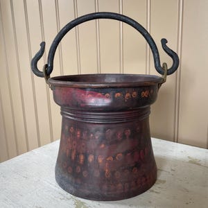 May include: A vintage copper bucket with a black handle and decorative accents. The bucket has a dark, mottled finish with reddish-brown and black tones. The handle is arched, and the bucket has a flared base.
